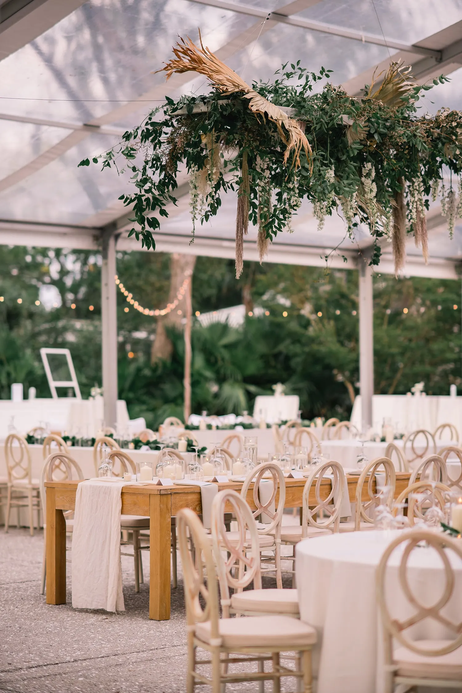 Reception under clear tent with greenery installation and wooden chairs