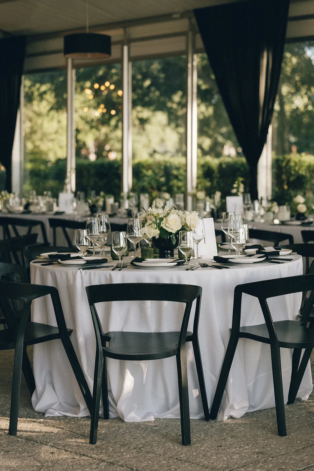 Round dining table with white linens, black chairs, and floral centerpiece by window.