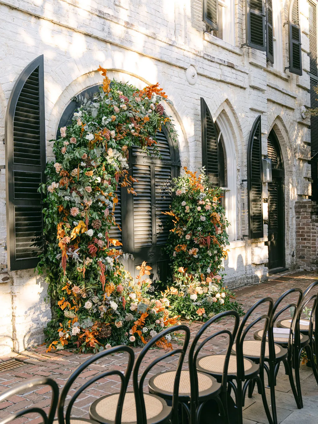 Floral installation on building exterior with chairs set for ceremony seating. | Curated Events Floral archway with vibrant autumn flowers and greenery against a brick wall with black shutters, set in a cozy outdoor event space with empty chairs, enhancing climate-conscious event design in Nashville.