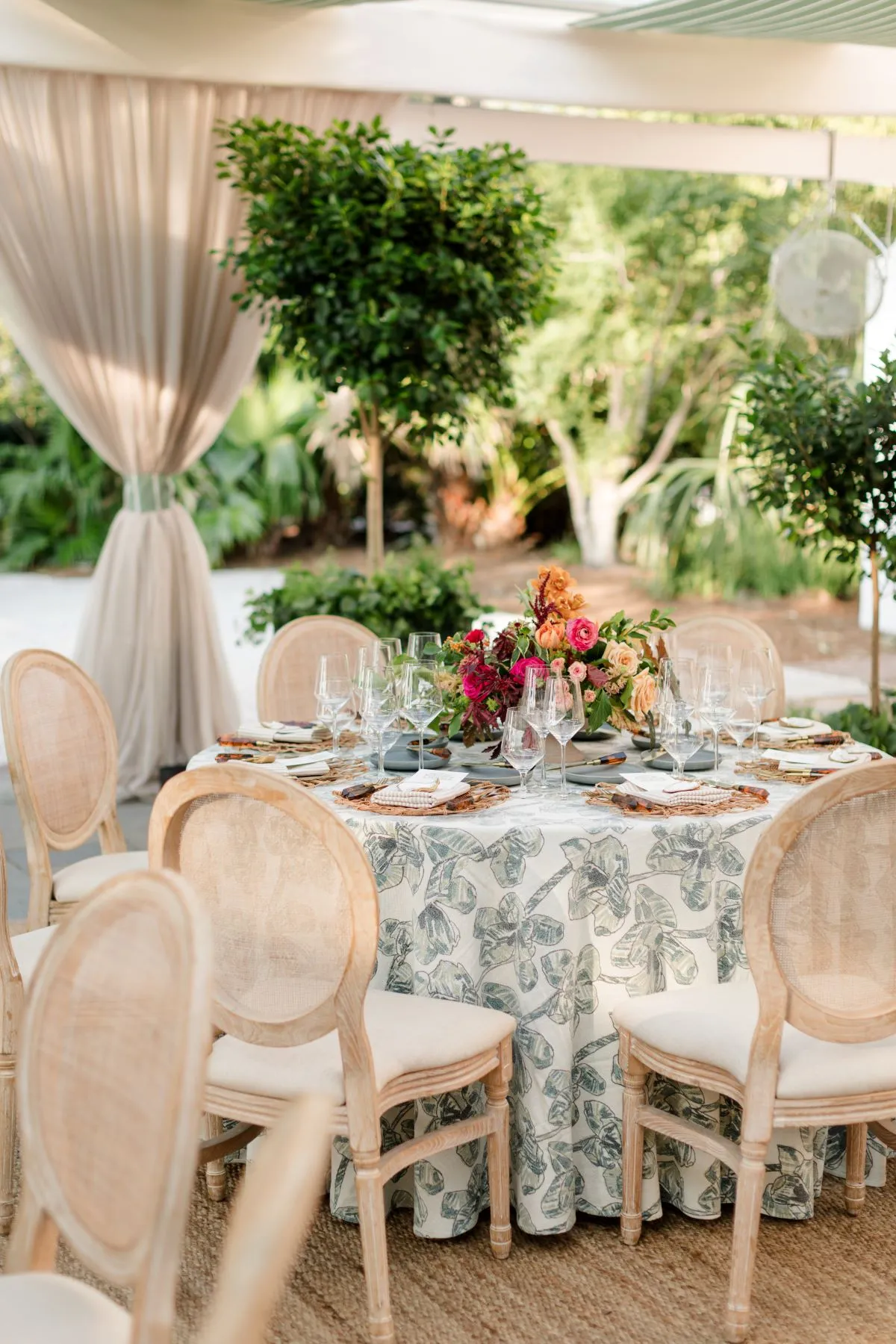 Round table with patterned linen, floral centerpiece, and upholstered chairs outdoors