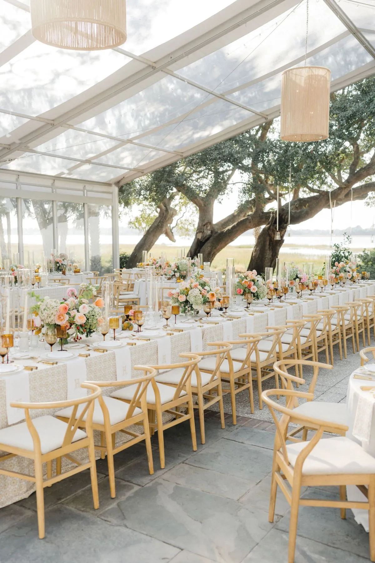 Long banquet table under clear tent with wooden chairs and lush floral centerpieces.