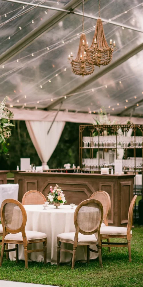 Tented outdoor bar and seating area with round table and wooden chairs for rainy event setup.