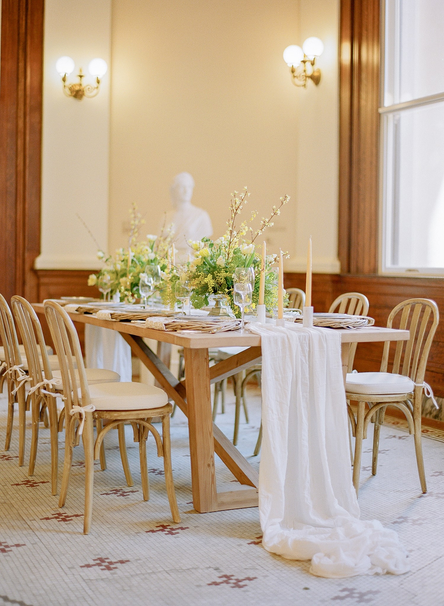Long wooden table set for a formal event with cream floral centerpiece, tall candles, and a white table runner in a sunlit room.