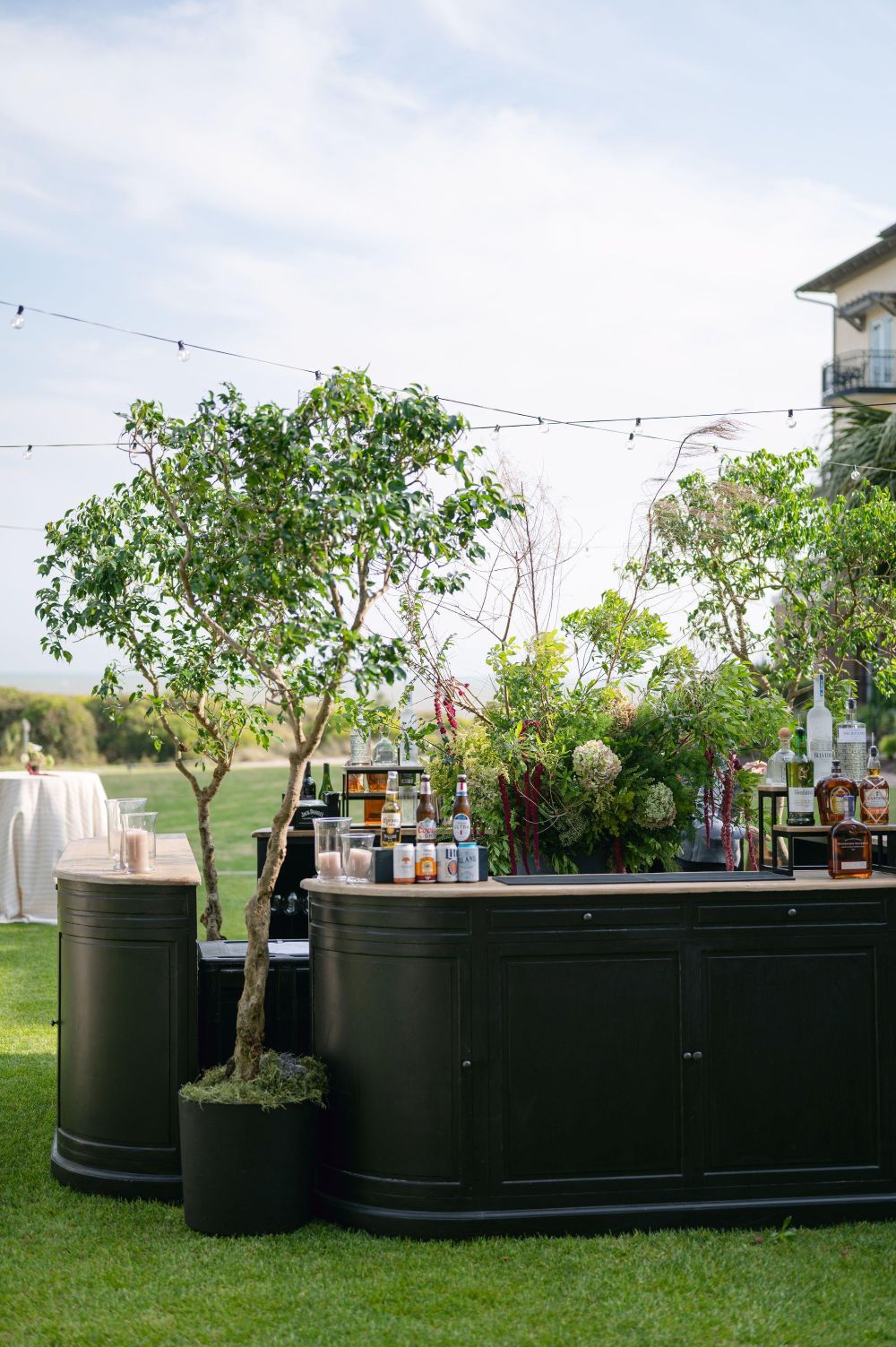 Outdoor bar setup with a curved black counter, bottles and greenery, on a grassy lawn under string lights.
