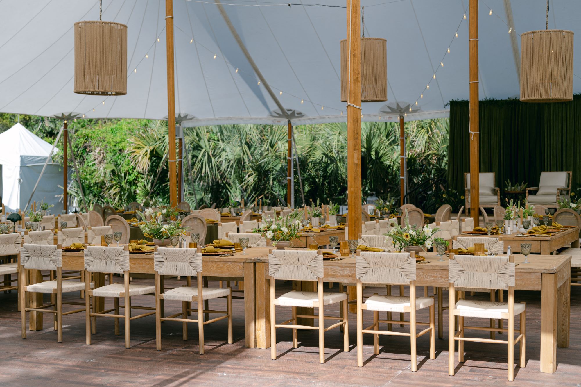 Long wooden dining tables set under a sailcloth tent with woven lighting and natural greenery accents | Curated Events Long wooden dining tables set under a sailcloth tent with woven lighting and natural greenery accents