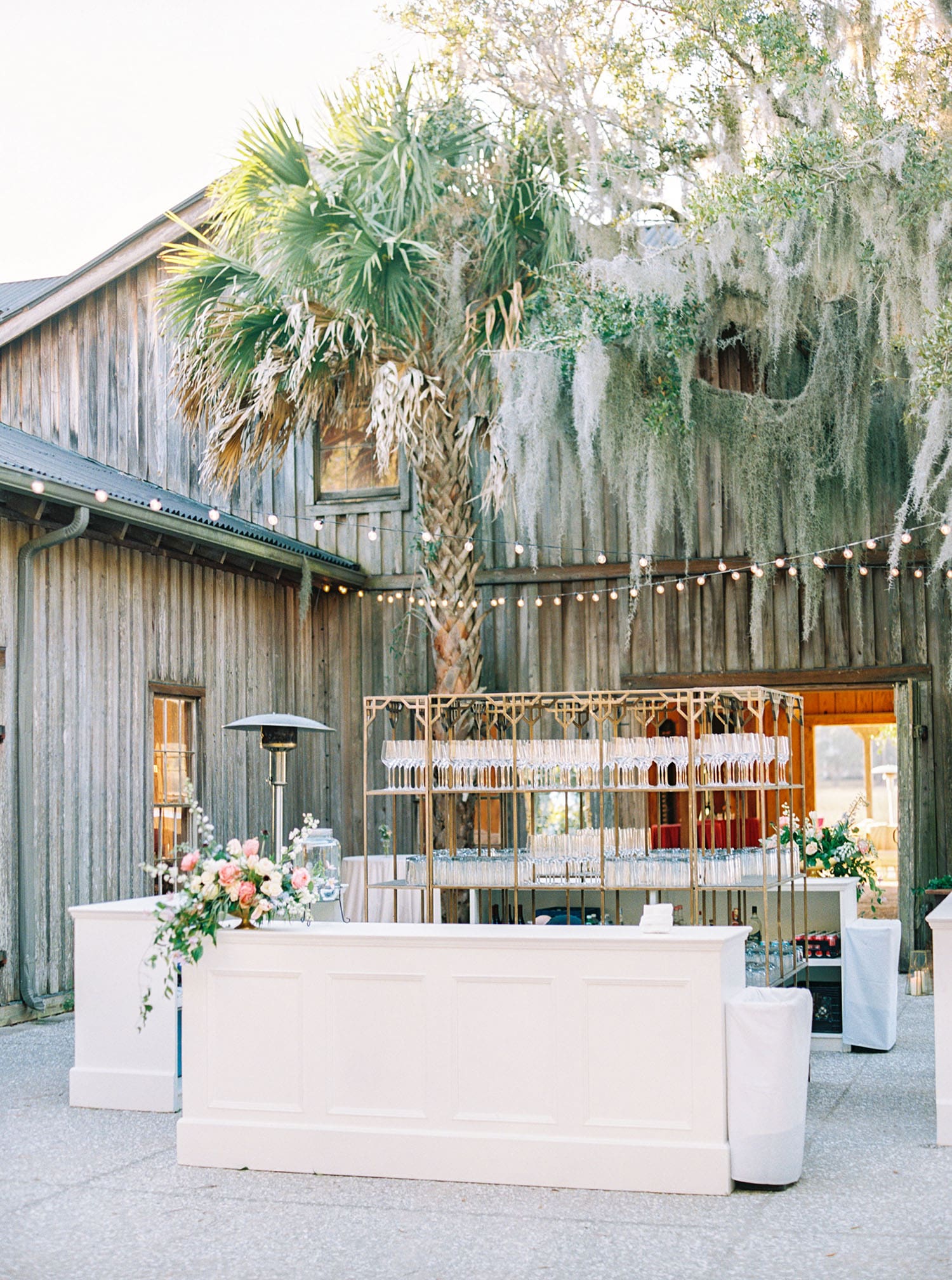 Corporate event bar installation in a rustic outdoor venue with glassware display and floral accents. | Curated Events Corporate event bar installation in a rustic outdoor venue with glassware display and floral accents.