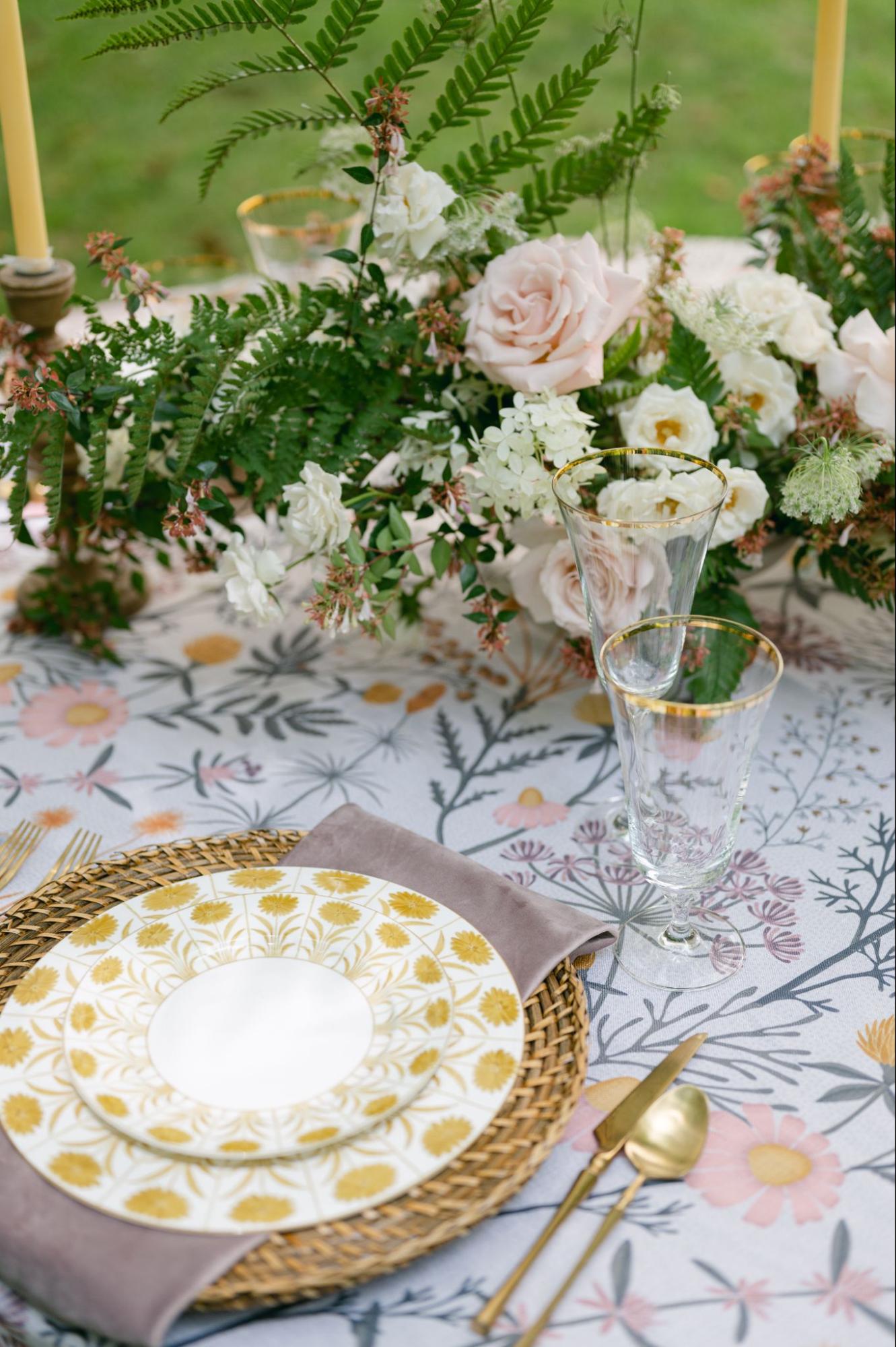 Floral summer place setting with woven charger, gold flatware, and patterned tabletop linens.