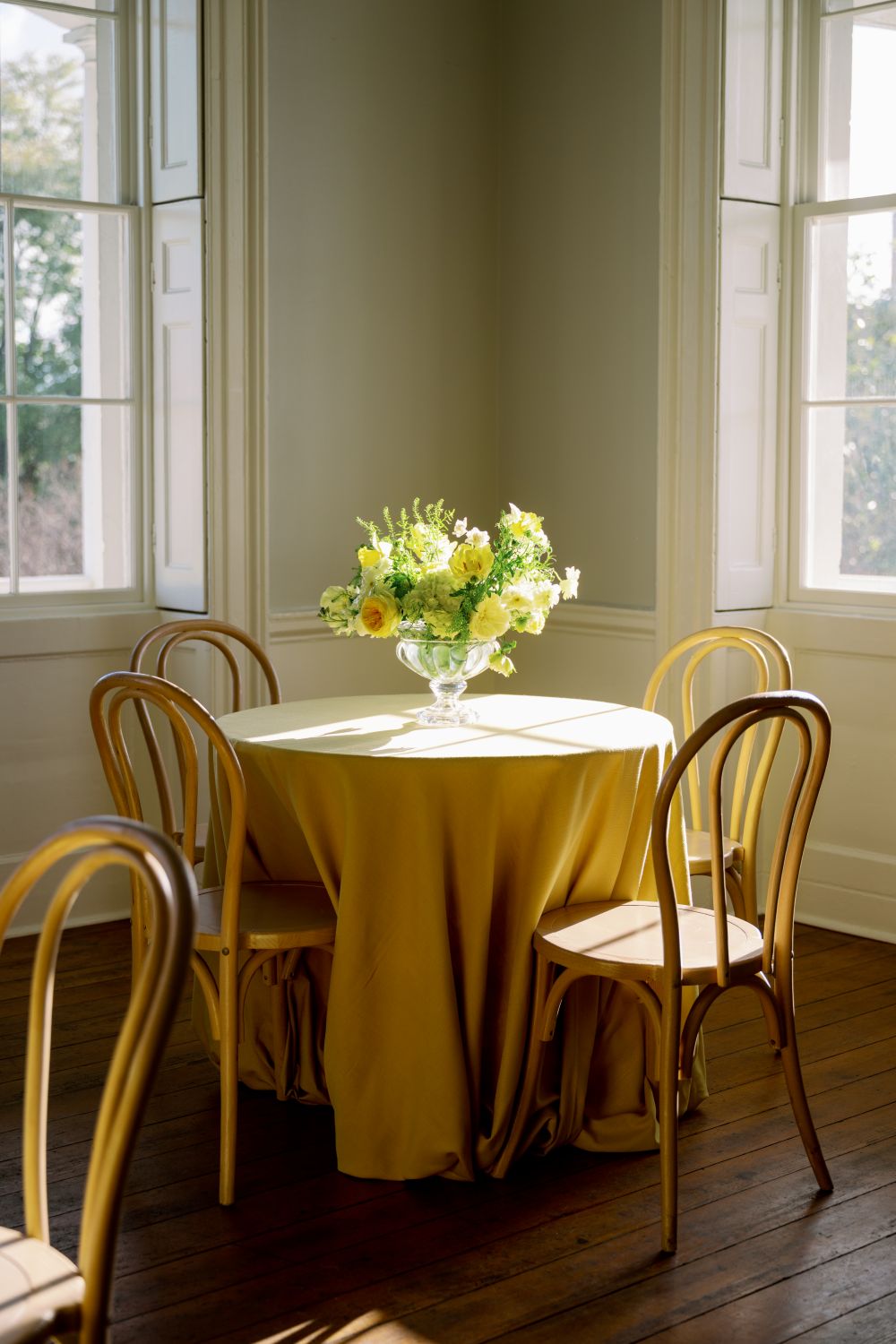 Round table with mustard linen, bentwood chairs, and yellow floral centerpiece by sunny windows.
