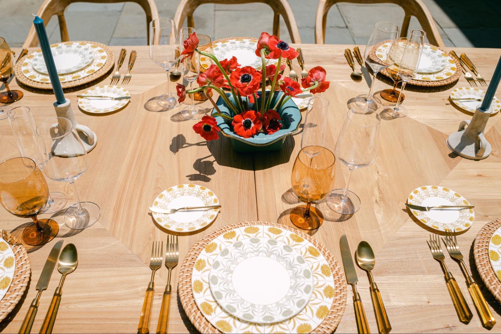 Sunny outdoor table with patterned plates, amber glassware, blue candles, and red florals. | Curated Events Sunny outdoor table with patterned plates, amber glassware, blue candles, and red florals.