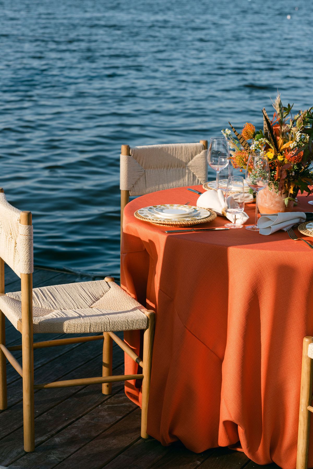 Waterfront table setting with rust linen, woven chairs, and warm floral centerpiece. | Curated Events Waterfront table setting with rust linen, woven chairs, and warm floral centerpiece.