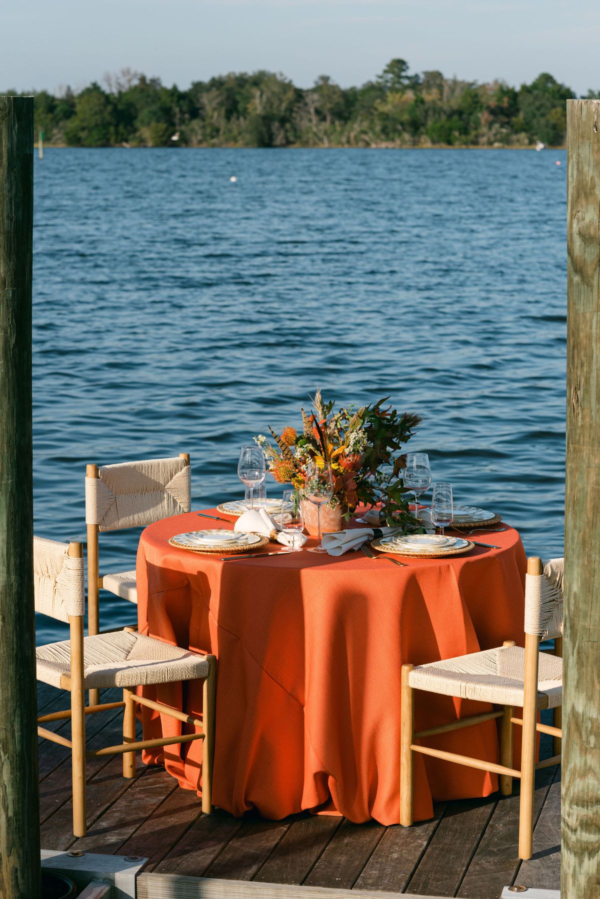 Waterfront event table with orange linen, wooden chairs, and floral centerpiece