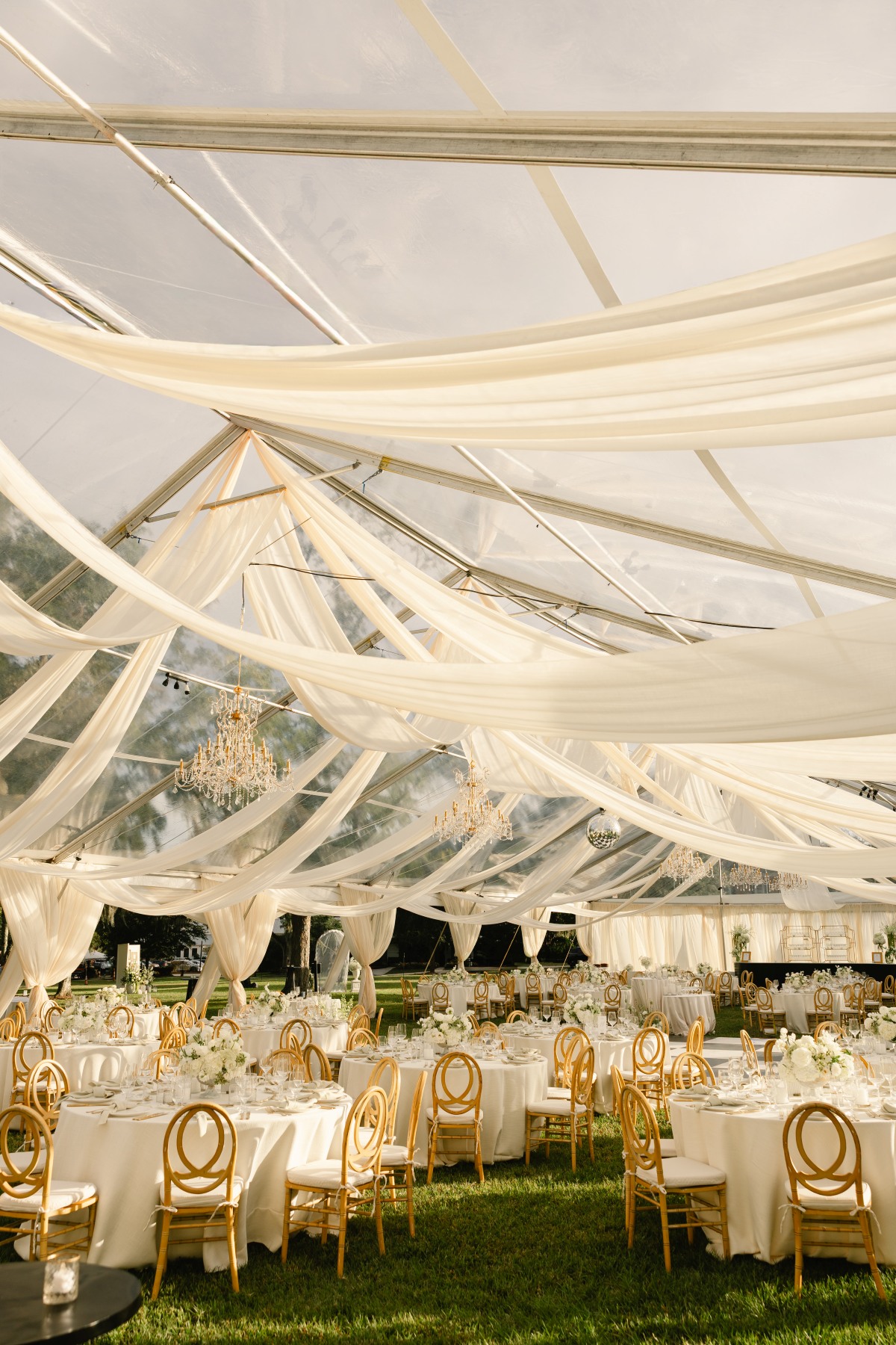 Outdoor summer event tent with sheer white draping, dining tables, and elegant rental seating | Curated Events Outdoor summer event tent with sheer white draping, dining tables, and elegant rental seating
