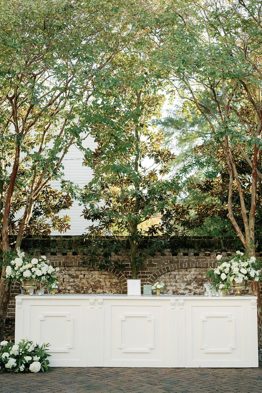 White paneled wedding bar framed by white flowers, mature trees, and a historic brick garden wall. | Curated Events White paneled wedding bar framed by white flowers, mature trees, and a historic brick garden wall.