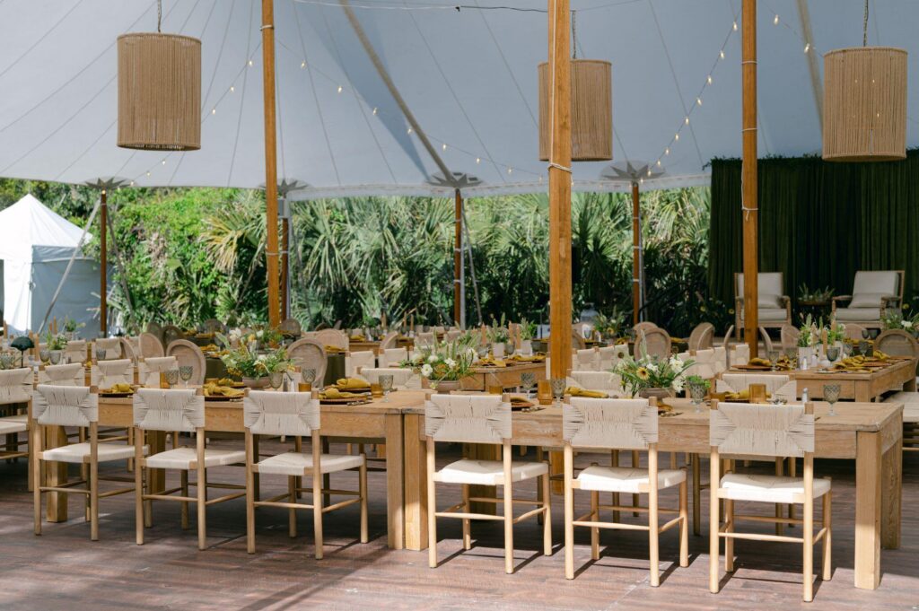 Elegant outdoor dining setup with wooden tables, chairs, and floral arrangements under a tent.