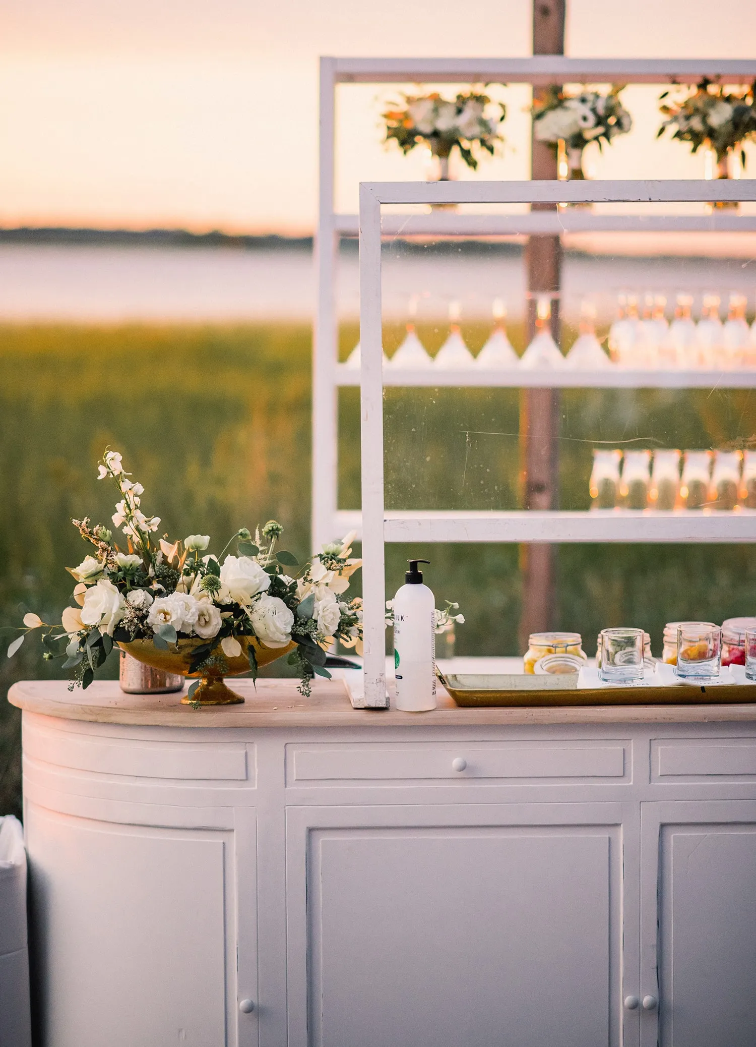Curved pale bar with open shelving, white flowers, and displayed glassware overlooking the water at sunset. | Curated Events Curved pale bar with open shelving, white flowers, and displayed glassware overlooking the water at sunset.
