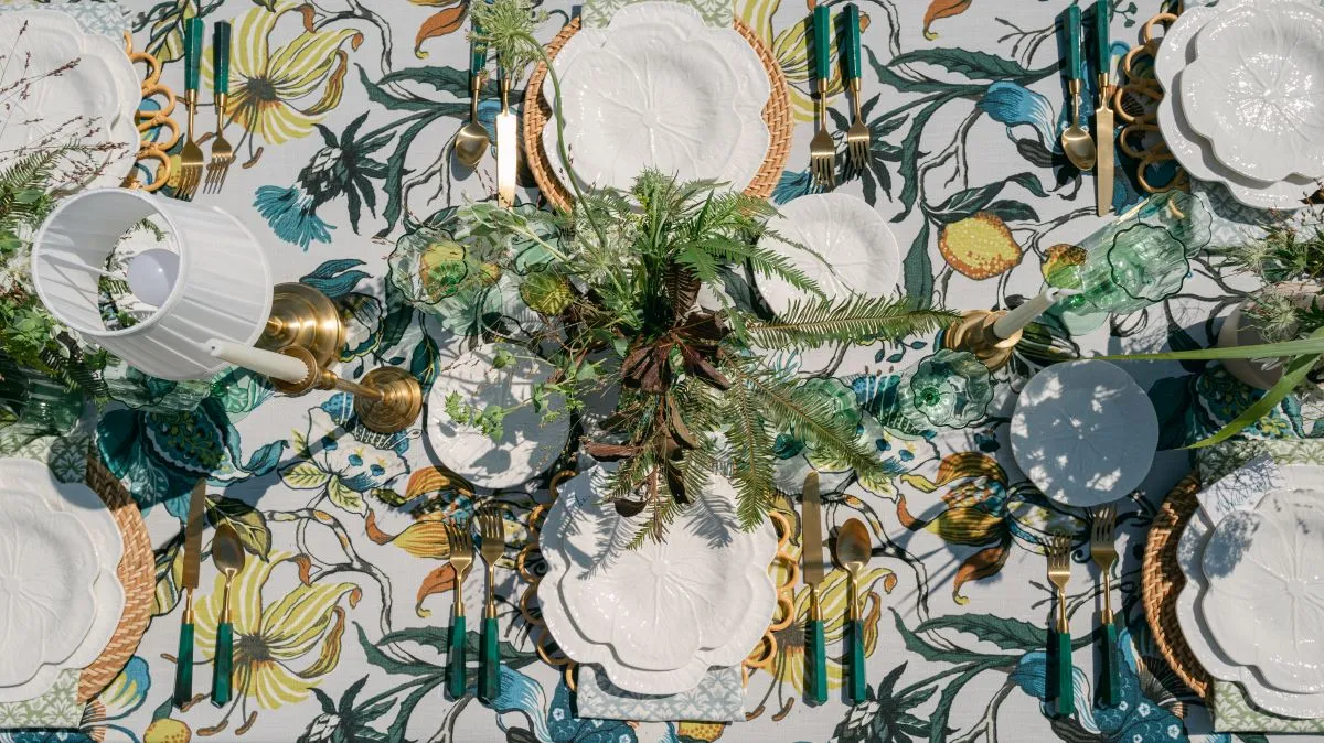 Overhead tablescape with botanical linen, white plates, green glassware, brass flatware, and natural greenery.