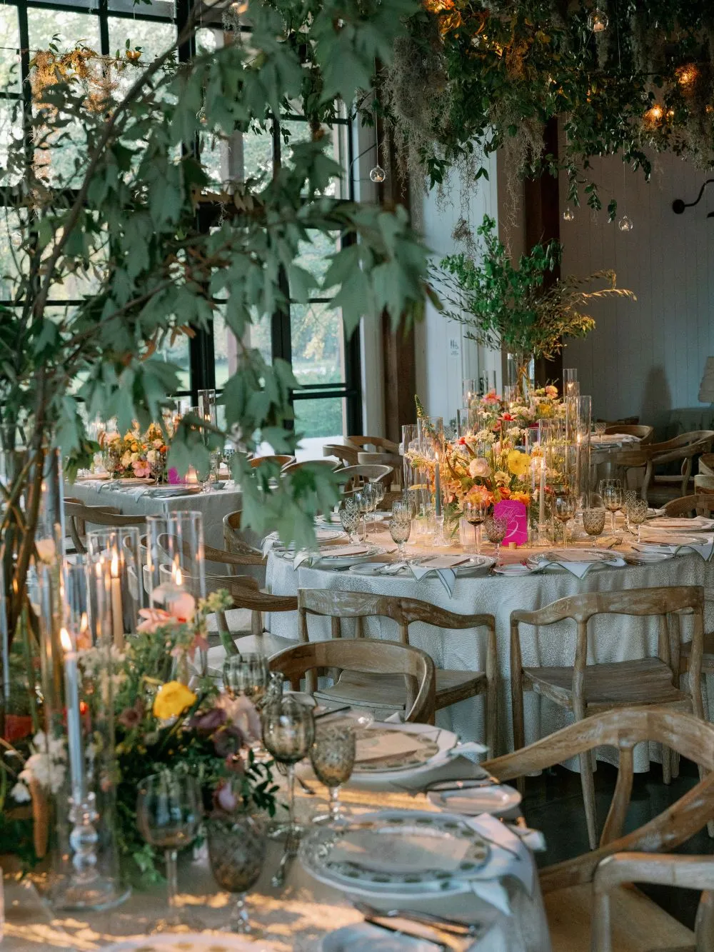 Indoor reception tables with patterned goblets, colorful flowers, glass candleholders, wooden chairs, and hanging greenery.
