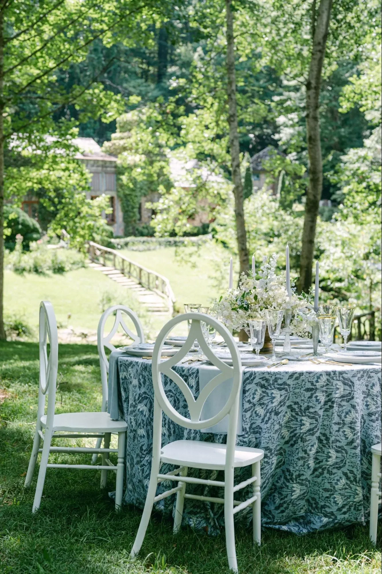Outdoor wedding table with blue botanical linen, white chairs, taper candles, and white flowers