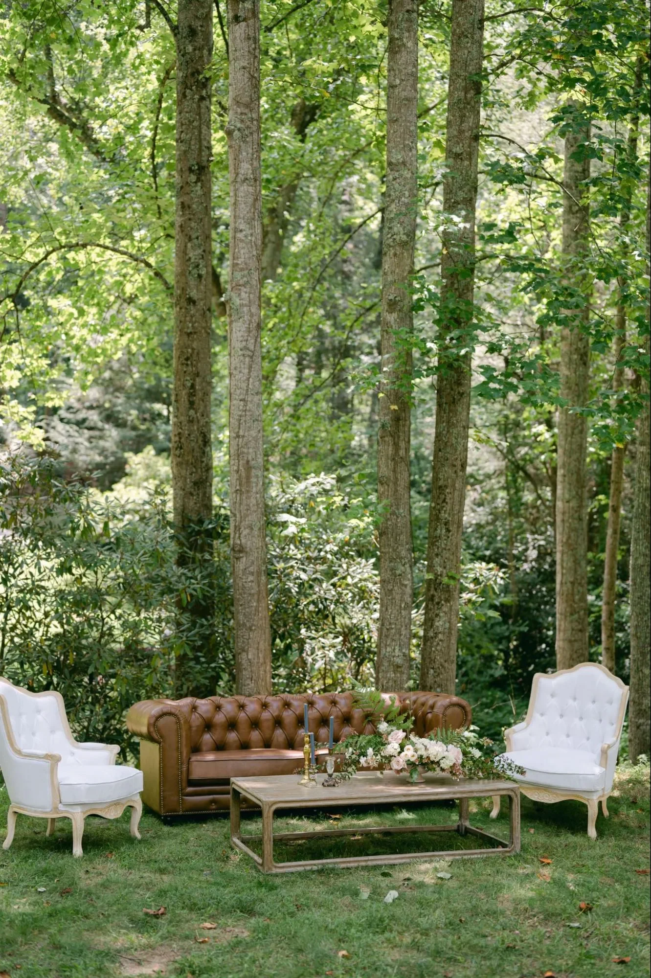 Brown leather sofa and white armchairs arranged as an outdoor woodland wedding lounge