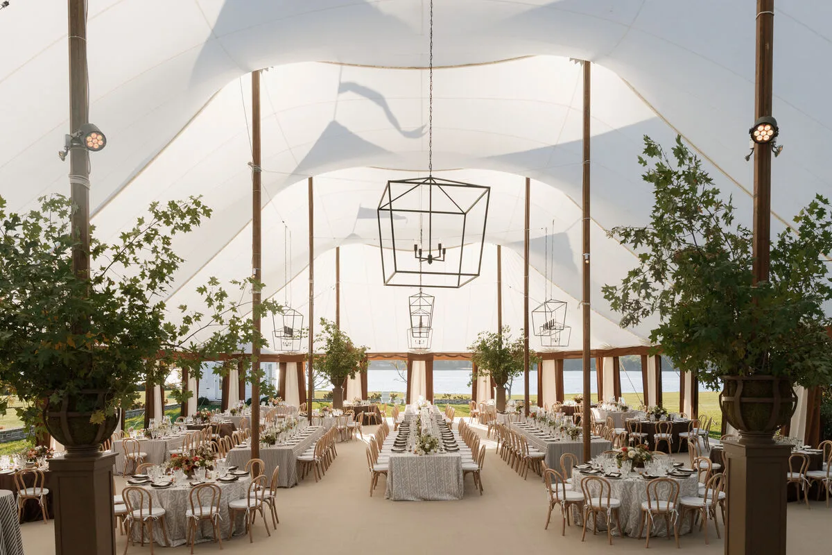 Sailcloth reception tent with round and long dining tables, large potted trees, and geometric chandeliers.