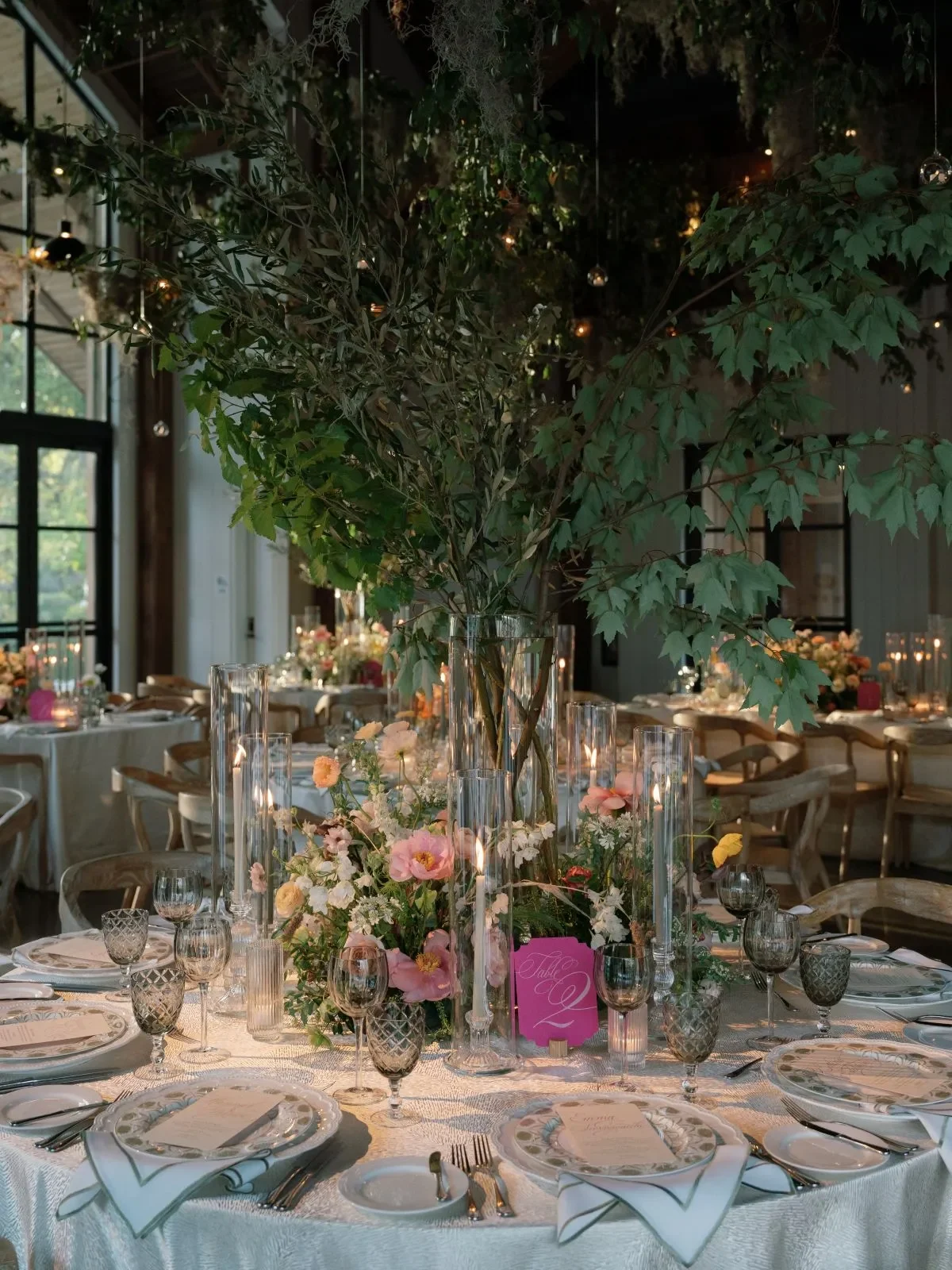 Round reception table with a tall greenery centerpiece, pink flowers, candles, and glassware Round reception table with a tall greenery centerpiece, pink flowers, candles, and glassware