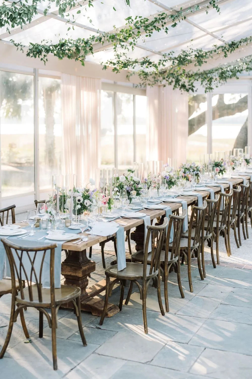 Long wood farm table styled with pale blue textiles, white florals, glass candleholders, and cross-back chairs beneath hanging greenery.