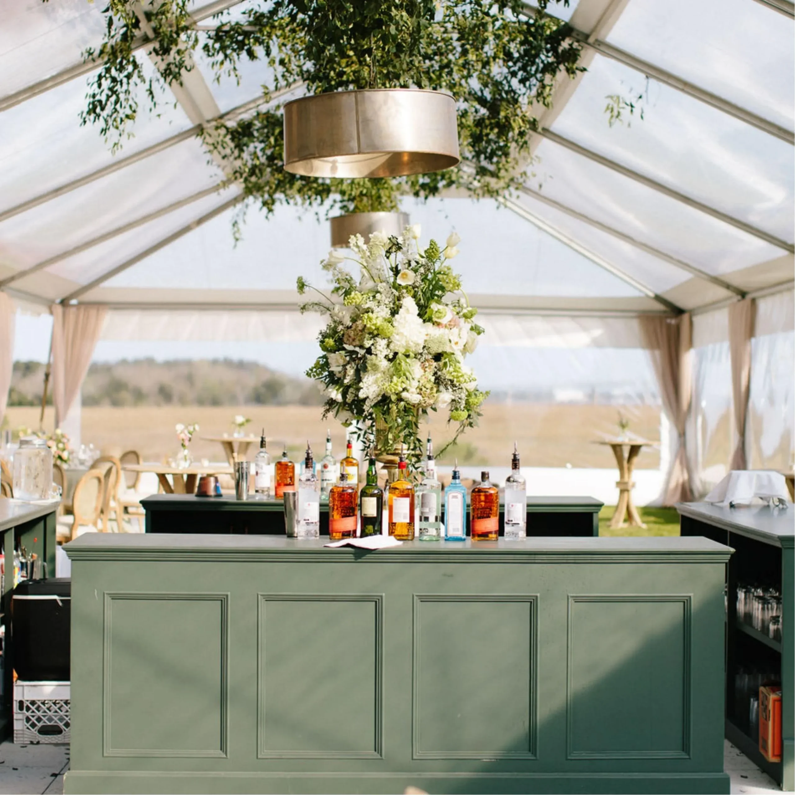 Green paneled bar with liquor bottles and a tall white floral arrangement inside a clear tent