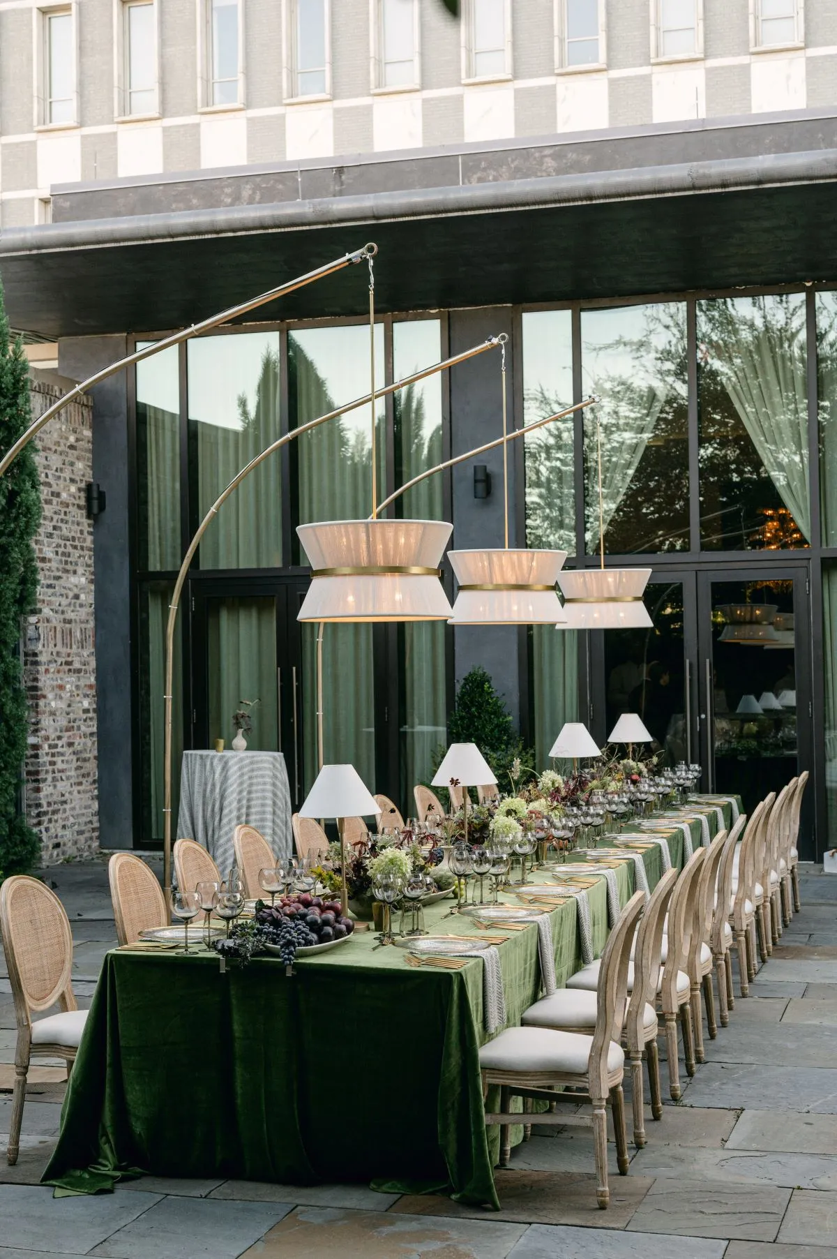 Long outdoor banquet table with neutral dining chairs, dark green linen and hanging lamps