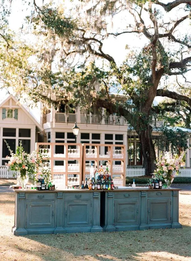 Blue-grey modular bar with wood back-bar shelving and floral arrangements beneath a large oak tree. | Curated Events Blue-grey modular bar with wood back-bar shelving and floral arrangements beneath a large oak tree.