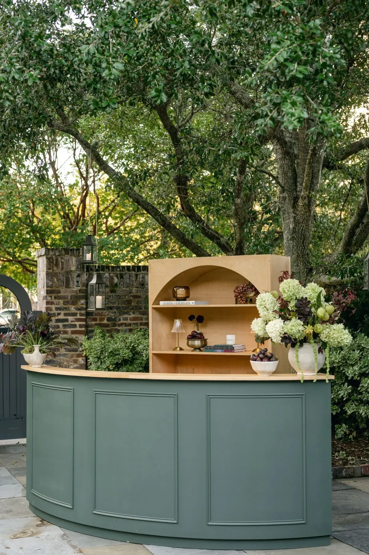 Curved sage green bar with wood shelving, fruit displays, flowers, and decorative lamps