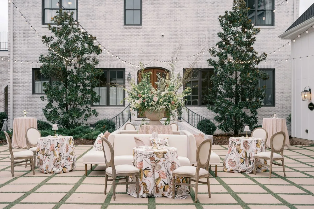 Courtyard lounge vignette with a curved neutral sofa, upholstered chairs, botanical-print tables, greenery, and a large floral arrangement.