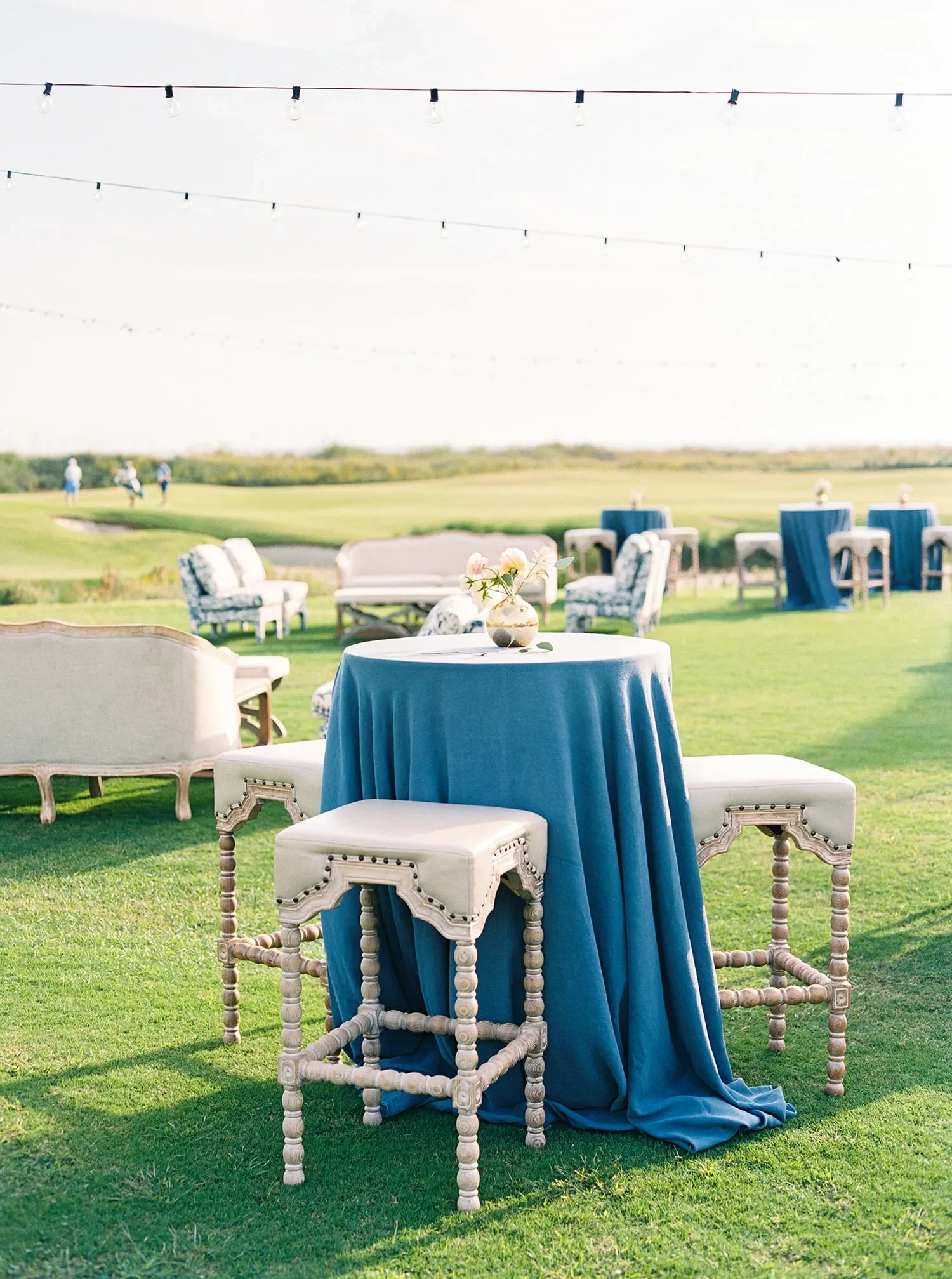 Cocktail table with deep blue linen and upholstered stools on an outdoor lawn