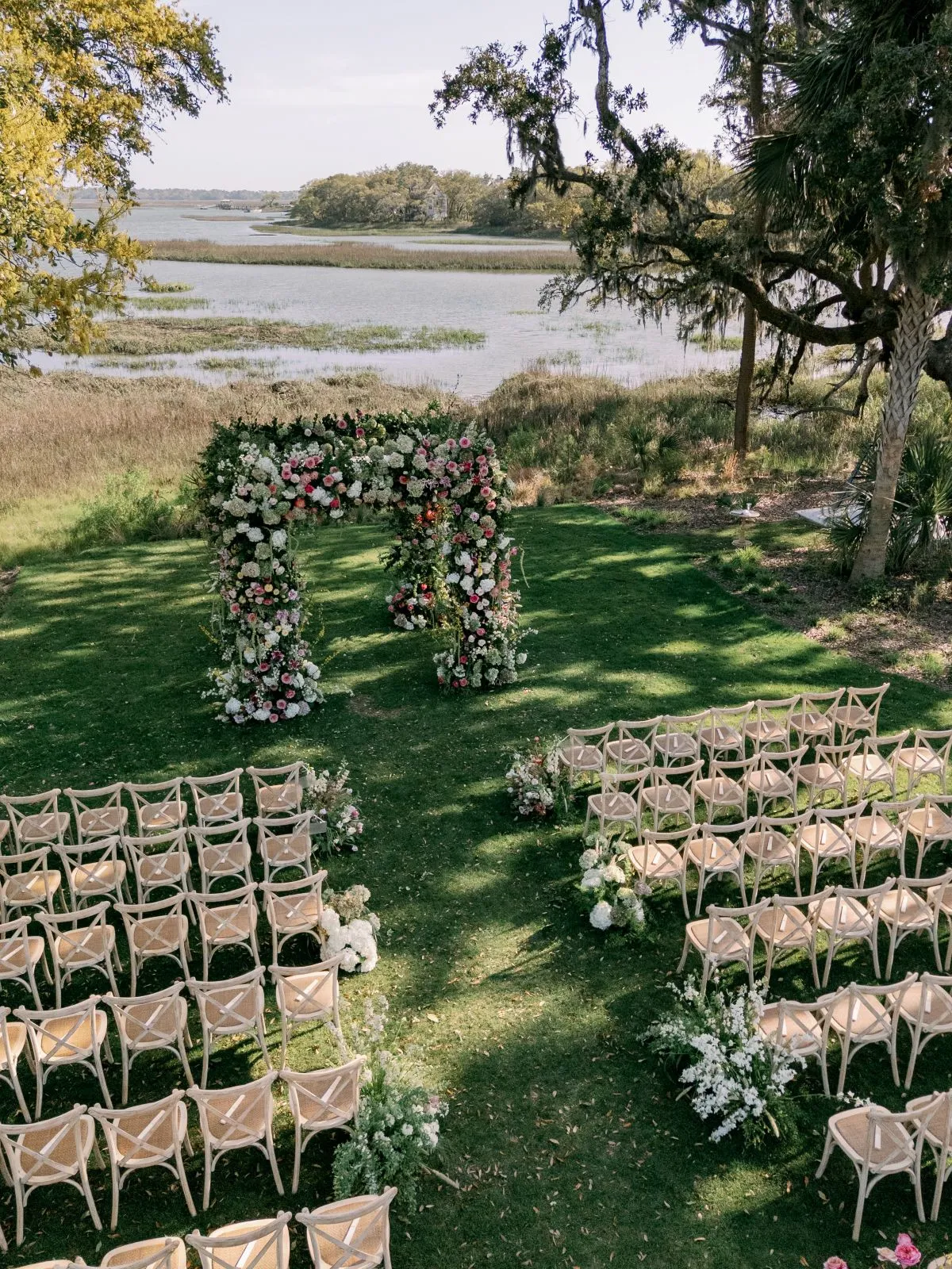 Outdoor Charleston ceremony with a lush floral arch, natural wood seating, aisle flowers, oak trees, and expansive marsh views. | Curated Events Outdoor Charleston ceremony with a lush floral arch, natural wood seating, aisle flowers, oak trees, and expansive marsh views.
