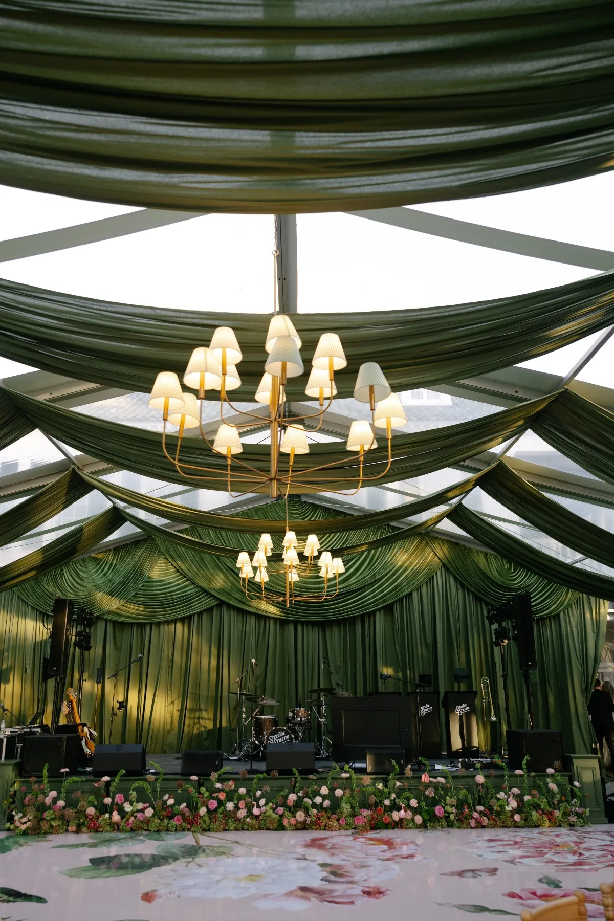 Event tent with dark green draping, shaded chandeliers, a floral-lined stage, and a painted dance floor.