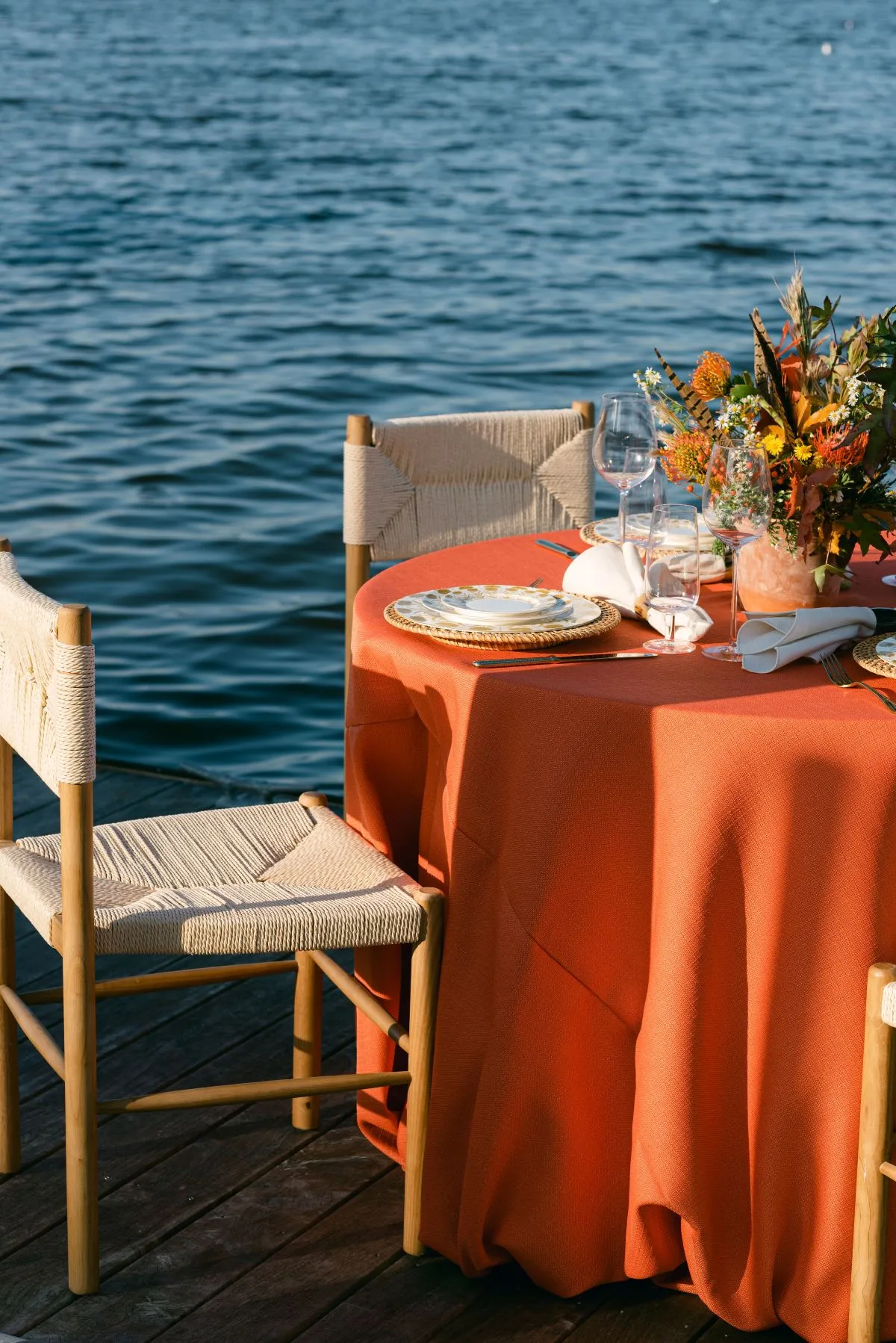 Waterfront dining table with orange linen, woven chairs, floral centerpiece, and glassware positioned beside the water.