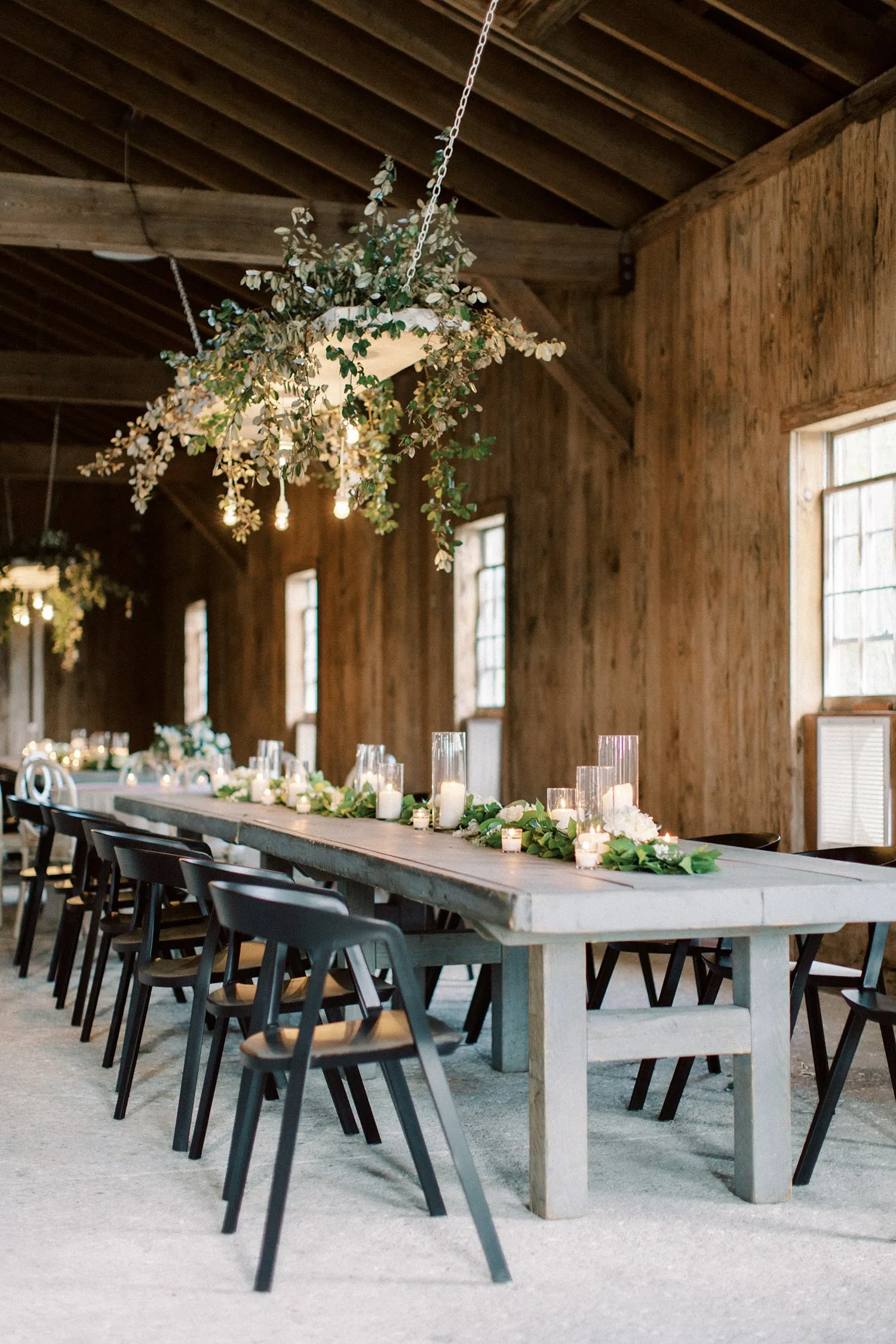 Long dining table with dark chairs, candles, greenery, and suspended floral lighting inside a rustic wood venue.