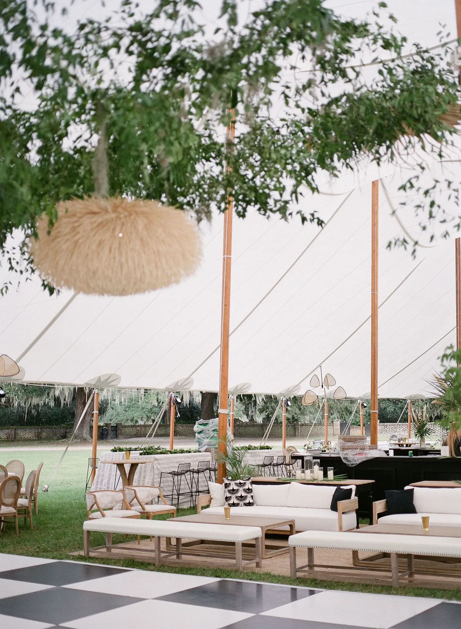 Open-sided wedding tent with a black-and-white dance floor, lounge seating, hanging greenery, and a woven pendant light.