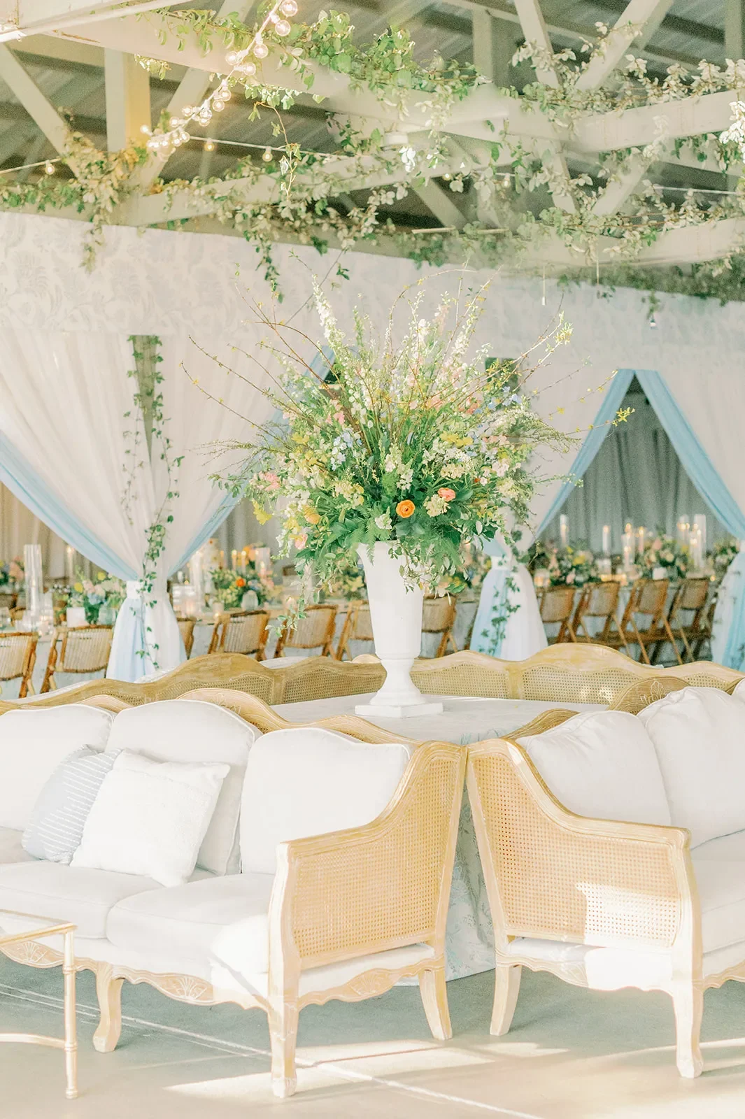 Cream cane lounge seating arranged around a tall floral display inside a wedding tent