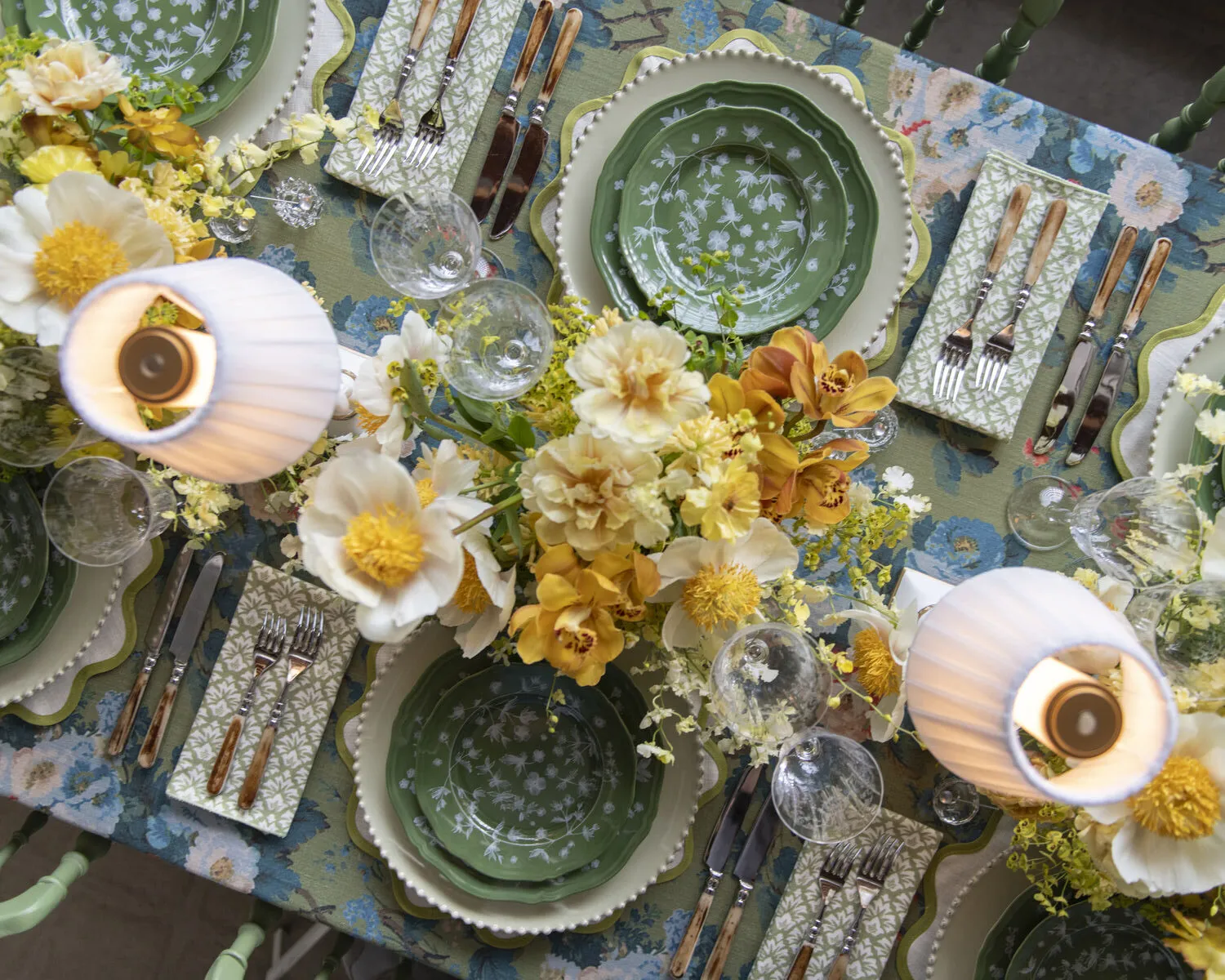 Overhead fall tablescape with botanical linen, green patterned china, white and ochre flowers, clear glassware, and shaded table lamps.