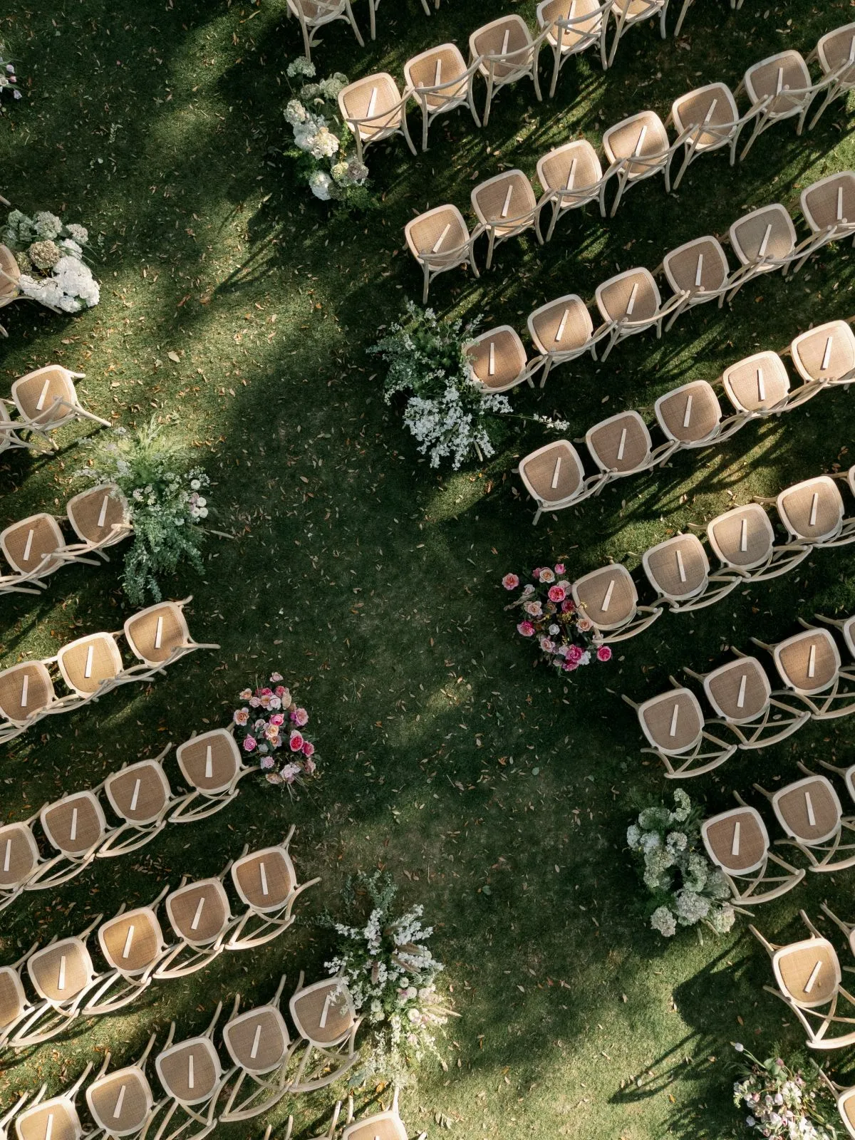 Overhead view of outdoor ceremony seating arranged around a central aisle with natural wood chairs and scattered floral arrangements. | Curated Events Overhead view of outdoor ceremony seating arranged around a central aisle with natural wood chairs and scattered floral arrangements.