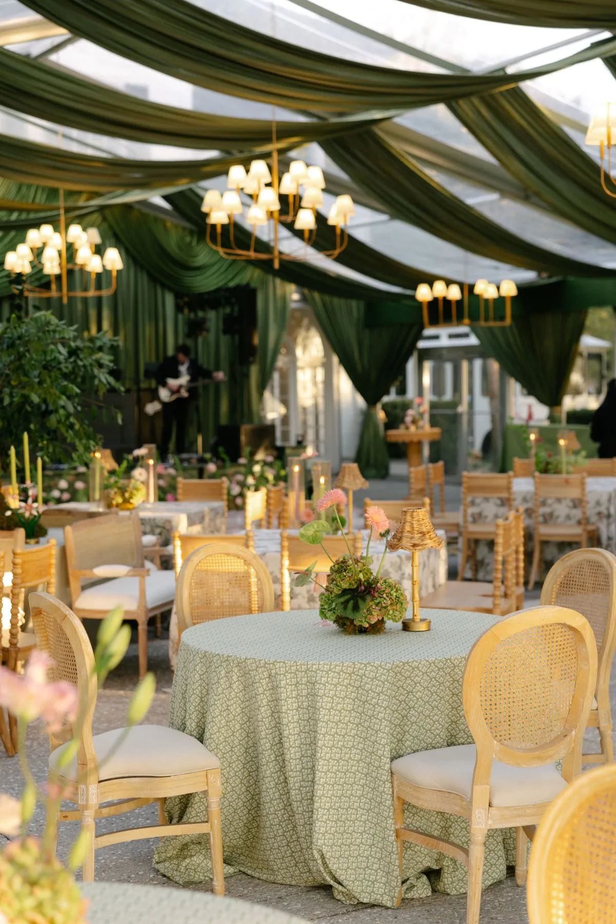 Cane-back chairs around pale green reception tables beneath dark green tent draping and gold chandeliers. | Curated Events Cane-back chairs around pale green reception tables beneath dark green tent draping and gold chandeliers.