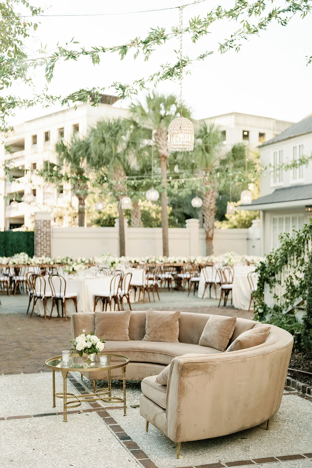 Charleston courtyard lounge with curved neutral sofas, gold tables, dining tables, palm trees, and suspended greenery overhead.