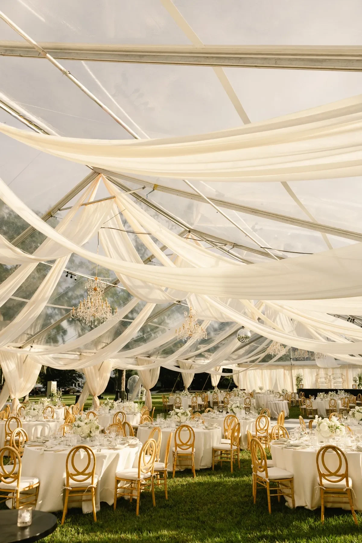 Clear wedding tent with ivory draping, crystal chandeliers, round tables, and gold chairs