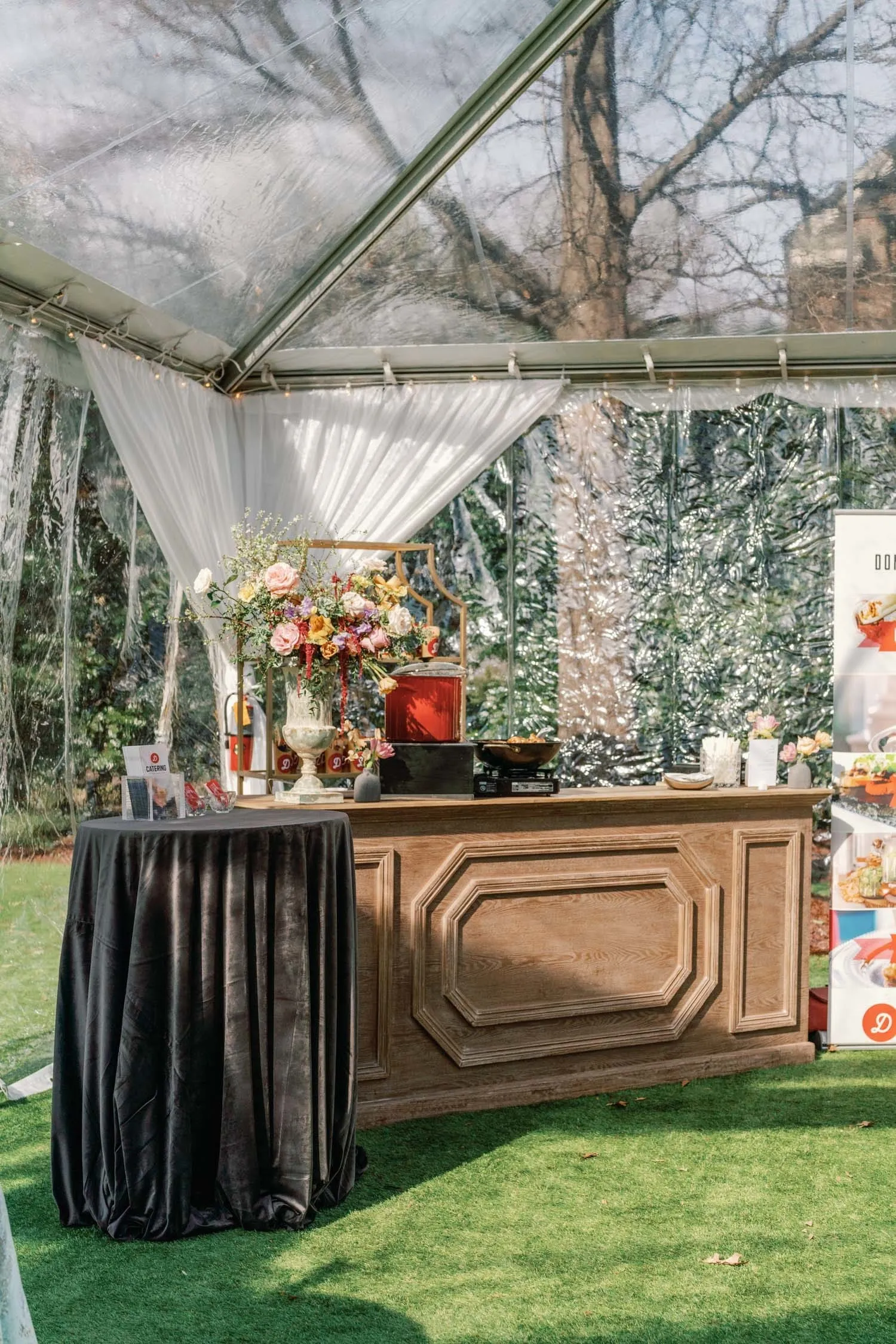 Carved wood bar under a clear tent with a black cocktail table and colorful floral arrangement