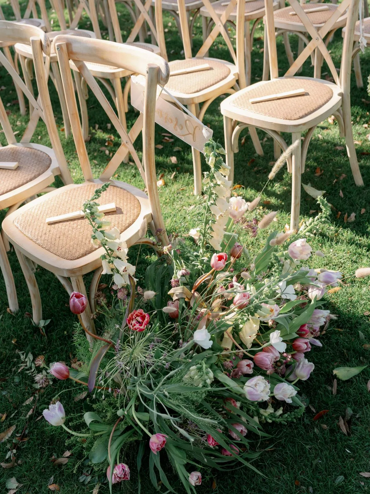 Close-up of natural wood ceremony chairs with woven seats and loose pink, white, and green florals arranged along the aisle. | Curated Events Close-up of natural wood ceremony chairs with woven seats and loose pink, white, and green florals arranged along the aisle.