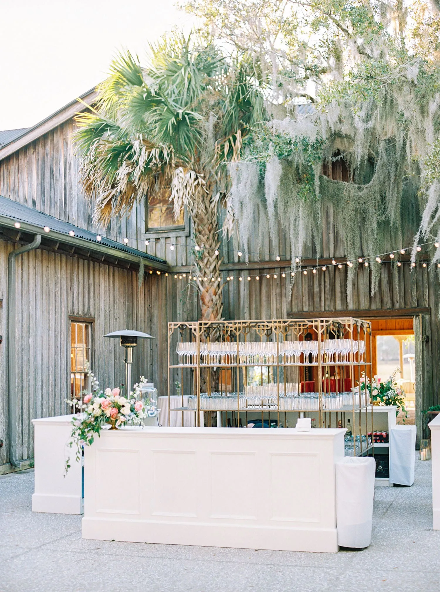 White event bar with brass shelving and floral accents at a rustic outdoor venue
