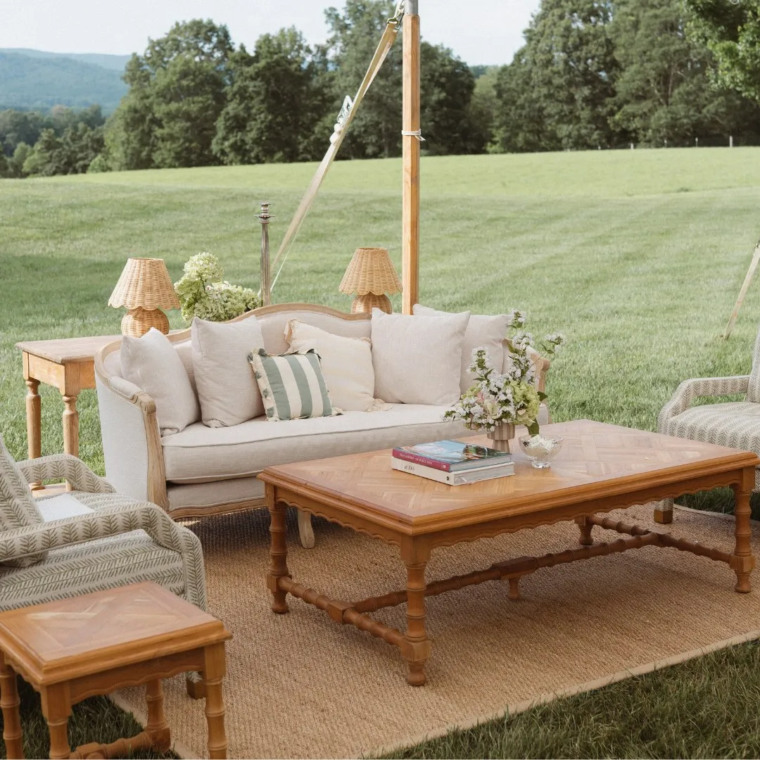 Vintage-inspired outdoor lounge with a neutral wood-frame sofa, woven chairs, wood tables, striped pillows, and small shaded lamps on a grassy field.