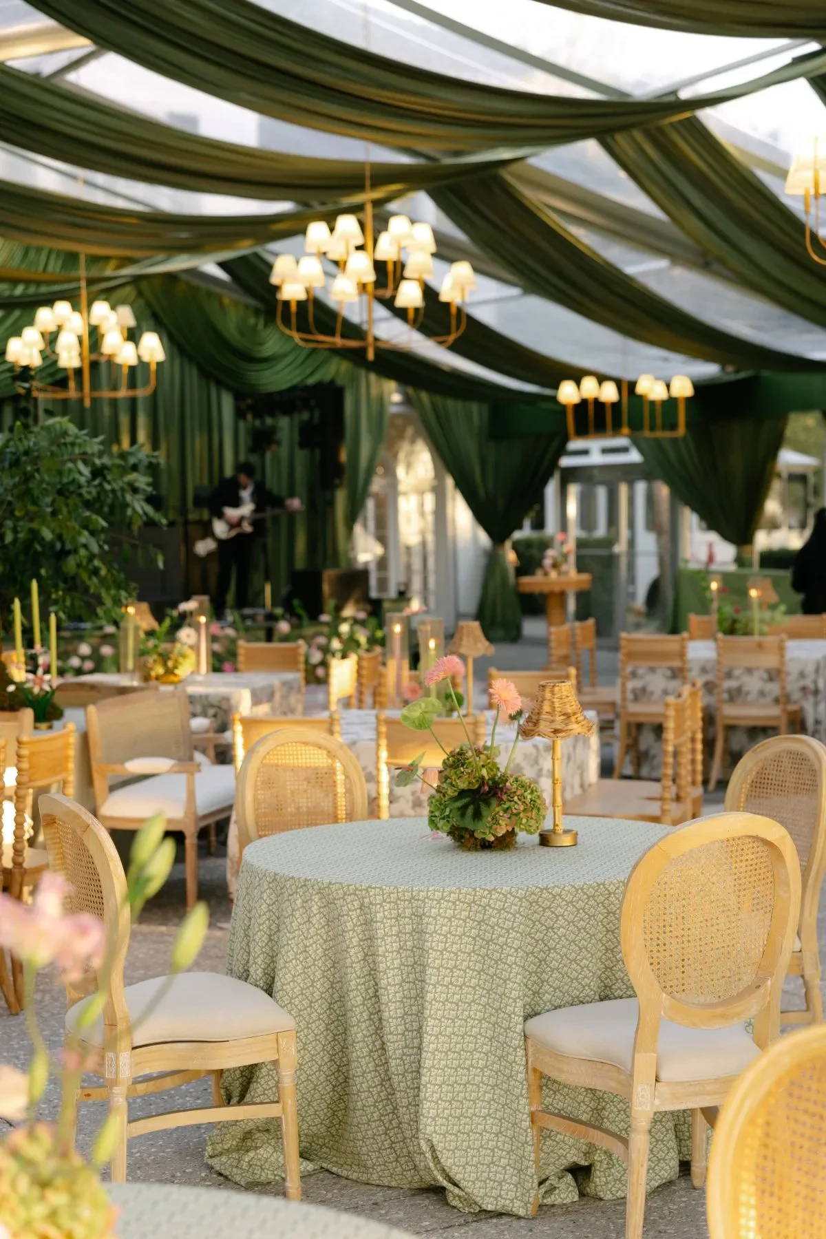 Round wedding table with sage patterned linen, cane chairs, flowers, and a small lamp Round wedding table with sage patterned linen, cane chairs, flowers, and a small lamp