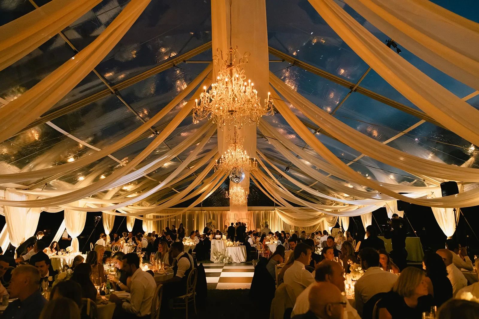 Evening wedding reception inside a clear-top tent with chandeliers and ivory ceiling draping