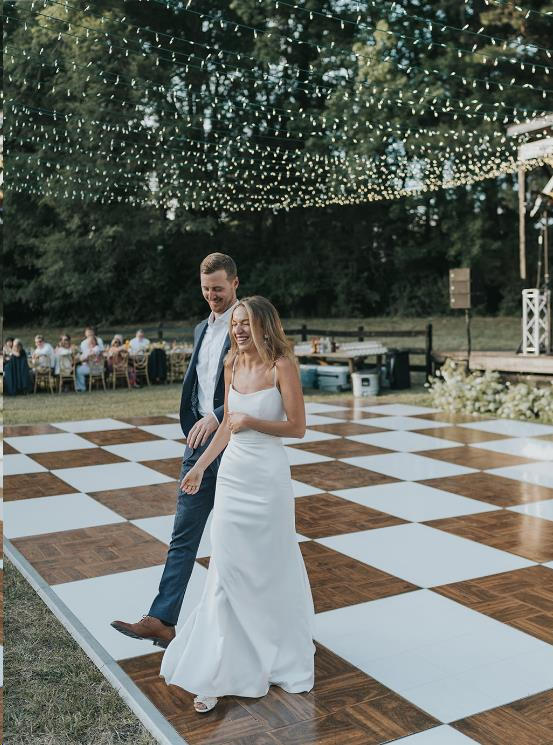 Bride and groom crossing a wood-and-white outdoor dance floor beneath string lights with a stage nearby.